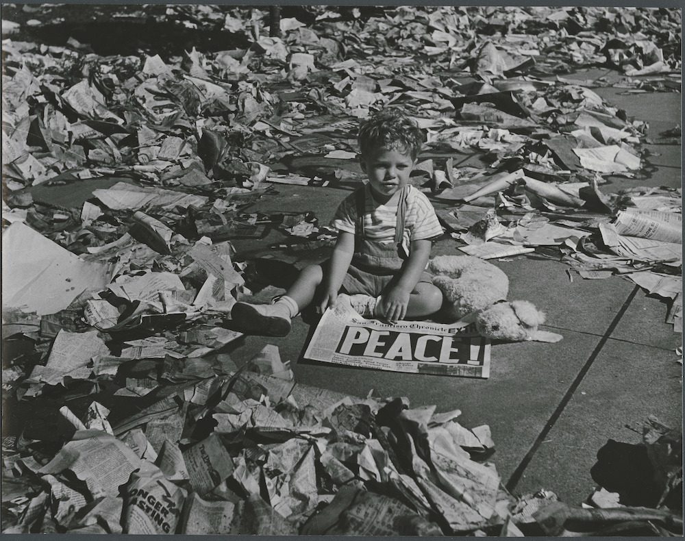young boy sitting on street amid litter, holding 'Peace' newspaper 1945 San Francisco