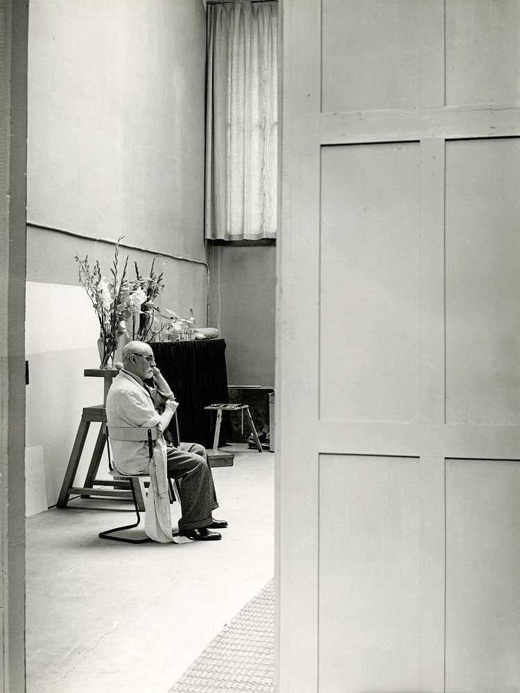 Picture with Matisse sitting in his studio and focused on something out of the photo shot. Photo by Brassaï (1939) - RM12690